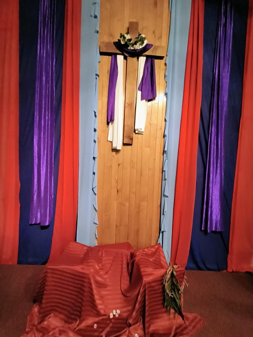 Wooden cross with colored fabric streamers of blue, red, and purple hanging vertically to the left and right sides